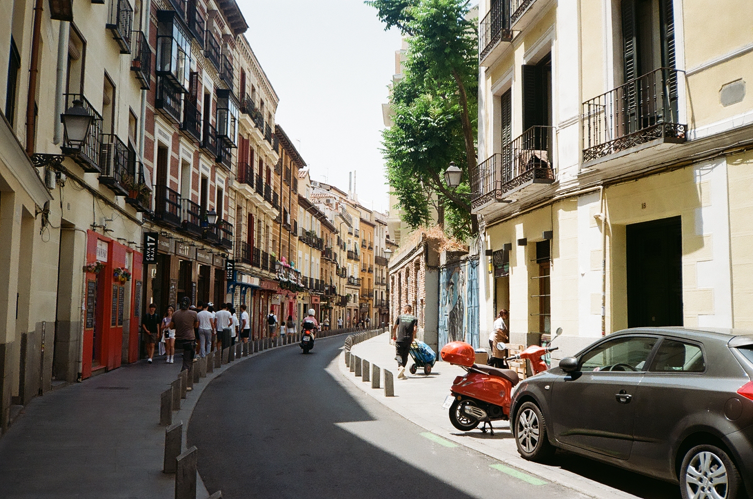 A narrow Spanish street with old-world architecture, a red Vespa, and pedestrians